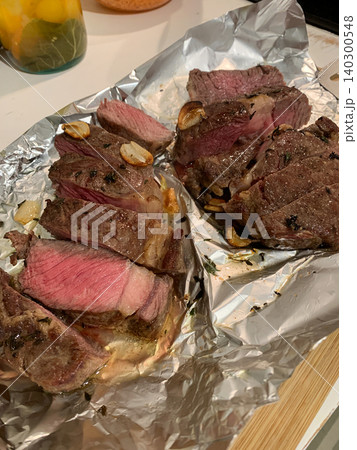 Steak is cut into portions and placed on foil with garlic. The scene shows a kitchen setting where food is being prepared for dinner. The evening light adds warmth to the scene Steak is cut into portions and placed on foil with garlic. The scene shows a kitchen setting where food is being prepared for dinner. The evening light adds warmth to the scene 140300548