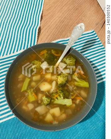 A bowl of potato and broccoli soup has chunks of carrots and onions. It sits on a blue and white striped tablecloth with a silver spoon beside it A bowl of potato and broccoli soup has chunks of carrots and onions. It sits on a blue and white striped tablecloth with a silver spoon beside it 140300552