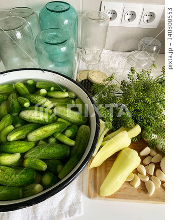 Cucumbers are soaking in a bowl while garlic and cucumber slices are on a cutting board. Several empty jars sit nearby on a clean countertop. It is a preparation activity for winter storage 140300553