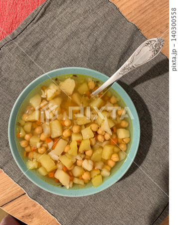 Bowl holds a warm potato and chickpea soup. The soup has chunks of potato and chickpeas in broth. A spoon rests in the bowl. Nearby is a glass filled with a drink 140300558