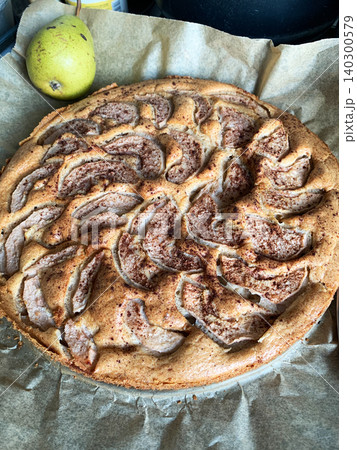 A gluten-free pear pie sits on a plate in a kitchen. The pie has slices of pear arranged on top. A fresh pear is beside the pie, creating a simple dessert scene in the afternoon light 140300579
