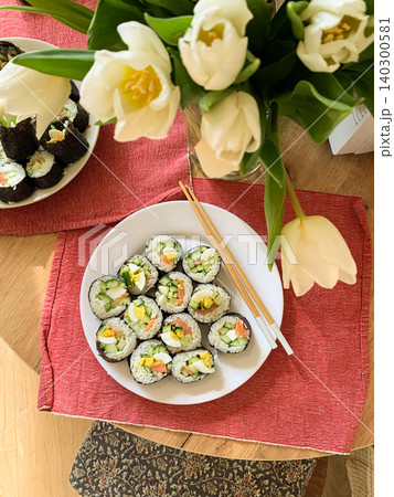 Sushi rolls are arranged neatly on a white plate. The rolls contain cucumber, red fish, and egg. They are served with chopsticks on a blue and green mat Sushi rolls are arranged neatly on a white plate. The rolls contain cucumber, red fish, and egg. They are served with chopsticks on a blue and green mat 140300581