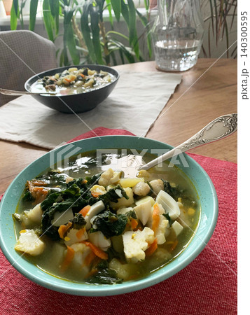 A bowl of nettle and egg soup is placed on a red cloth. The soup contains potatoes, cauliflower, and chopped greens. A spoon rests beside the bowl ready for use A bowl of nettle and egg soup is placed on a red cloth. The soup contains potatoes, cauliflower, and chopped greens. A spoon rests beside the bowl ready for use 140300595