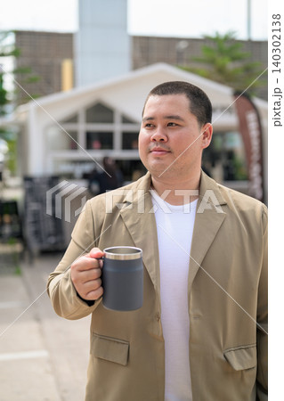 Plus size Isan Thai man holding a travel mug outdoors at a cafe 140302138