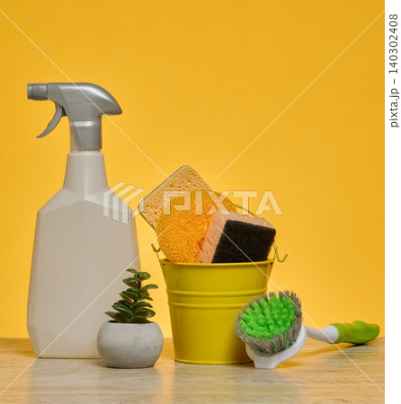 White plastic spray bottle and a stack of multi-colored sponges stand on a wooden surface against a yellow background. 140302408