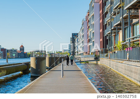 Amsterdam, Netherlands, 22.04.2026 - Modern residential buildings and waterfront promenade along zaan river with people walking on sunny day, contemporary dutch architecture Amsterdam, Netherlands, 22.04.2026 - Modern residential buildings and waterfront promenade along zaan river with people walking on sunny day, contemporary dutch architecture 140302758