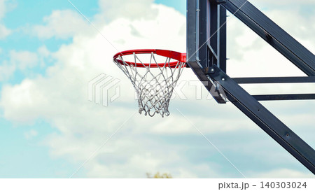 Outdoor basketball hoop with red rim against a blue sky with white clouds. 140303024