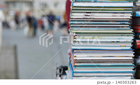 Stack of colorful books laying on a street vendor's stall with blurred people walking in the background 140303263
