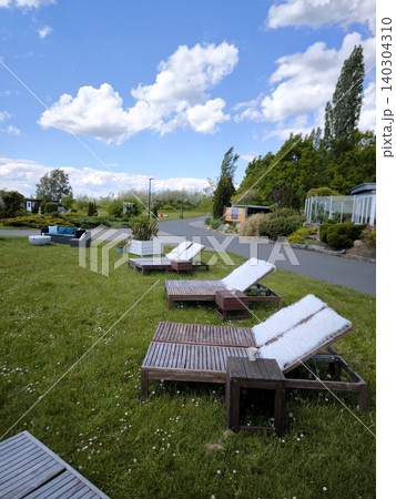 Wooden sun loungers, covered with cozy white throws, stand on the grass by the path. Wooden sun loungers, covered with cozy white throws, stand on the grass by the path. 140304310