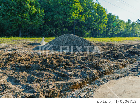 Construction site with gravel pile and cleared earth near wooded edge in sunlight 140306715