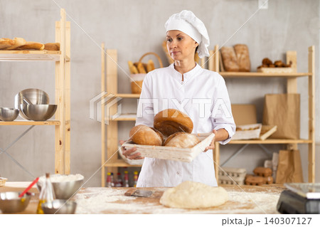 Young woman professional baker in apron standing at work table, posing with baked bread in bakery kitchen 140307127