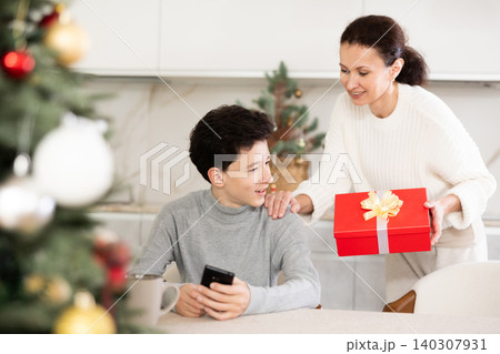Smiling mother giving box with Christmas gift to teenage son near New Year tree Smiling mother giving box with Christmas gift to teenage son near New Year tree 140307931
