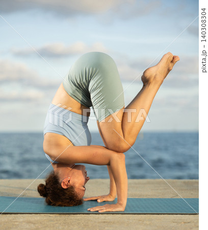 Sporty woman practitioner of yoga performing headstand, maintaining poised control and showcasing flexibility 140308304