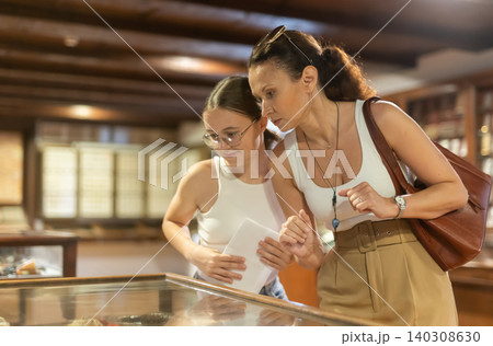 Young woman with teen daughter visitors stands near horizontal glass display case Young woman with teen daughter visitors stands near horizontal glass display case 140308630
