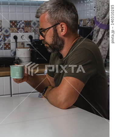 Man enjoys a cup of coffee while sitting at his kitchen table Man enjoys a cup of coffee while sitting at his kitchen table 140310285