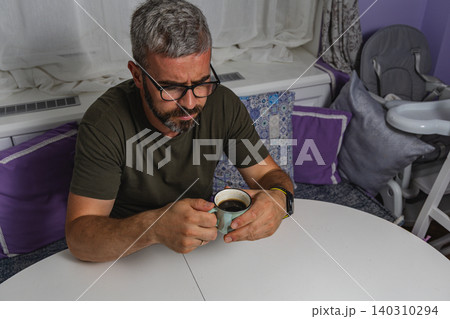 Man enjoys a hot cup of coffee while seated at a table in his cozy home Man enjoys a hot cup of coffee while seated at a table in his cozy home 140310294