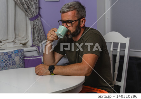 Adult man enjoys hot coffee while sitting at table in home interior Adult man enjoys hot coffee while sitting at table in home interior 140310296