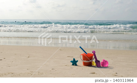 Children's colorful beach sand toy set including star mold, shovel, bucket and rake placed on sandy shore with sea waves behind. Play concept. Copy space Children's colorful beach sand toy set including star mold, shovel, bucket and rake placed on sandy shore with sea waves behind. Play concept. Copy space 140311964