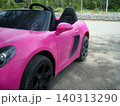 A side view of a bright pink toy convertible car parked on a paved path. The plastic electric vehicle features detailed black seats, a side mirror, dark rims, and a sporty profile 140313290
