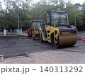 Two large yellow and orange road rollers sit on freshly laid black asphalt during street construction work. Traffic cones stand nearby, surrounded by green trees and buildings on a bright day 140313292