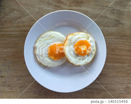 High angle view of two sunny-side-up fried eggs on a white round plate placed on a wooden table. High angle view of two sunny-side-up fried eggs on a white round plate placed on a wooden table. 140315450
