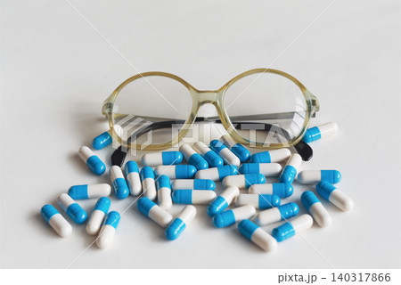 Macro shot of folded reading glasses lying among scattered white and blue capsules on a white background, for senior healthcare, medication management and vision support themes. Macro shot of folded reading glasses lying among scattered white and blue capsules on a white background, for senior healthcare, medication management and vision support themes. 140317866