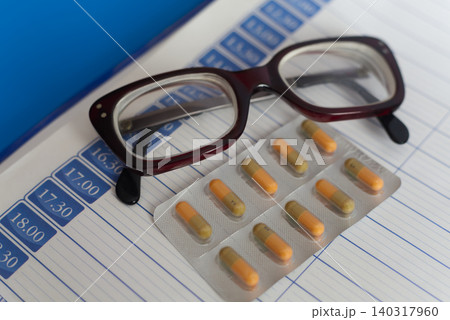 Close-up of reading glasses and a set of pills on a page of a paper daily planner, symbolizing treatment regimen for elderly people.  140317960