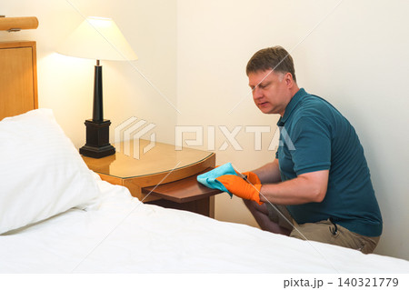 A man of European appearance cleans dust and dirt from a bedside table A man of European appearance cleans dust and dirt from a bedside table 140321779