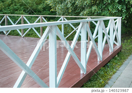 Outdoor summer terrace with brown wooden floor and railings in the form of a lattice against a background of green trees, close-up Outdoor summer terrace with brown wooden floor and railings in the form of a lattice against a background of green trees, close-up 140323987
