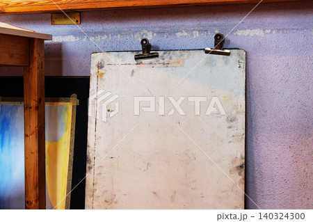 Used vertical drawing board with visible stains and clip holders, positioned against a textured purple wall in an art studio. Used vertical drawing board with visible stains and clip holders, positioned against a textured purple wall in an art studio. 140324300