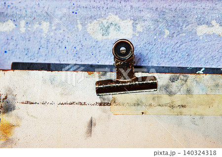 Macro view of a rusty metal clip attached to an aged and stained sketch board against a worn and textured purple wall. Macro view of a rusty metal clip attached to an aged and stained sketch board against a worn and textured purple wall. 140324318