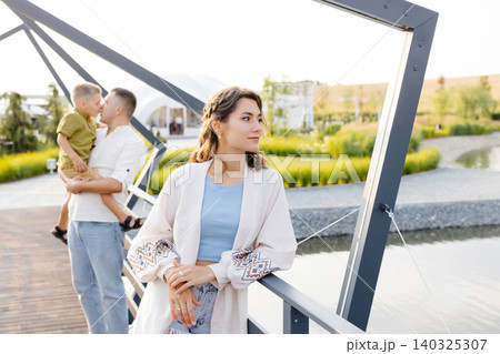 A Woman Dressed in a Flowing White Gown Stands Elegantly on a Picturesque Bridge, With Her Loving Husband Gently Holding Their Son by Her Side 140325307