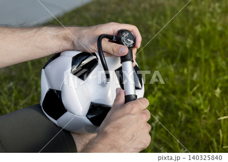 A man inflates a soccer ball with a hand pump featuring a pressure gauge, capturing a simple ritual of game preparation. A man inflates a soccer ball with a hand pump featuring a pressure gauge, capturing a simple ritual of game preparation. 140325840