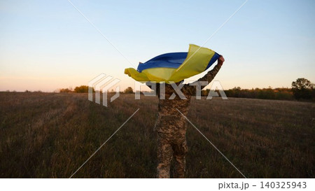 Soldier of ukrainian army running with raised flag Ukraine above his head at field. Young male military in uniform jogging with blue-yellow banner at meadow. Victory against russian aggression 140325943