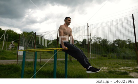 Strong man trains abdominal muscles raising legs on parallel bars at sports ground. Young shirtless sportsman exercising at nature. Sporty athlete working out at nature. Concept of sportive lifestyle Strong man trains abdominal muscles raising legs on parallel bars at sports ground. Young shirtless sportsman exercising at nature. Sporty athlete working out at nature. Concept of sportive lifestyle 140325968