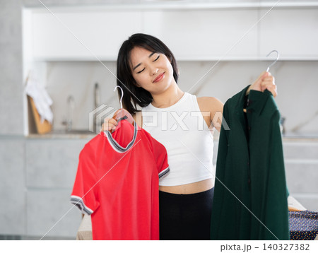 Happy young Asian girl trying to choose a dress in the kitchen Happy young Asian girl trying to choose a dress in the kitchen 140327382