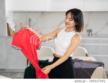 Happy young Asian girl trying to choose a dress in the kitchen 140327391
