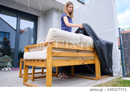 Woman handles black protective cover on wooden patio sofa with cushions outside home Woman handles black protective cover on wooden patio sofa with cushions outside home 140327731