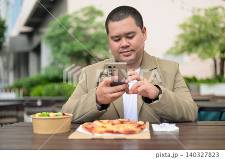 Focused plus size Isan Thai businessman texting near pizza outdoors 140327823