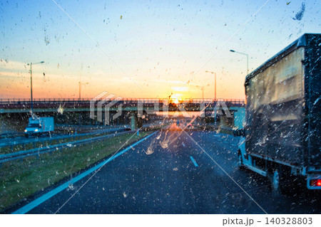 Highway traffic during sunset with trucks and cars through a dirty windshield, showcasing transportation, logistics, road safety and the challenges of visibility while driving on long journeys. Highway traffic during sunset with trucks and cars through a dirty windshield, showcasing transportation, logistics, road safety and the challenges of visibility while driving on long journeys. 140328803