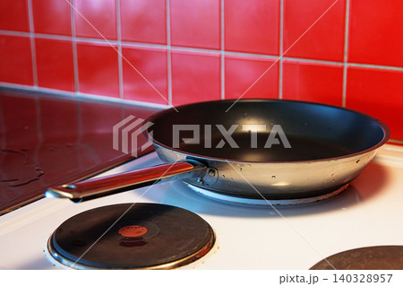 Coseup of a freshly washed clean teflon pan placed on a black cooking plate of a white electric stove with red tile backsplash. Coseup of a freshly washed clean teflon pan placed on a black cooking plate of a white electric stove with red tile backsplash. 140328957