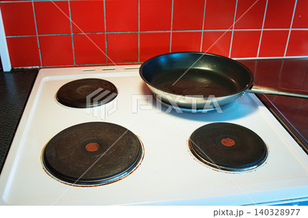 Black nonstick frying pan placed on a white electric stove with four round hotplates in a domestic kitchen with the vibant red tiled backsplash 140328977