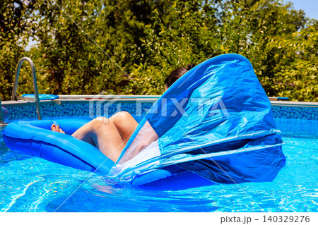 Female enjoys their time on blue inflatable lounger in swimming pool under sun with green plants nearby. Female enjoys their time on blue inflatable lounger in swimming pool under sun with green plants nearby. 140329276