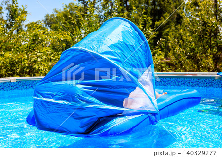 Female enjoys time on blue lounge float in pool with canopy in bright sun warm day. 140329277