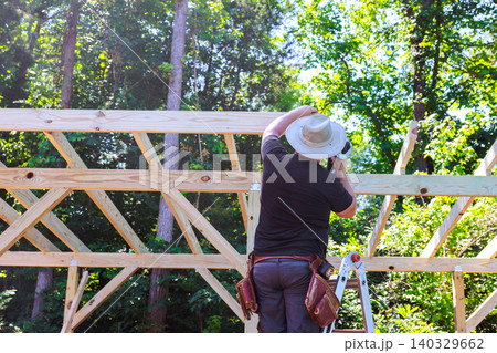 Worker uses tools on wooden beams joins structure they stand on ladder under trees Worker uses tools on wooden beams joins structure they stand on ladder under trees 140329662