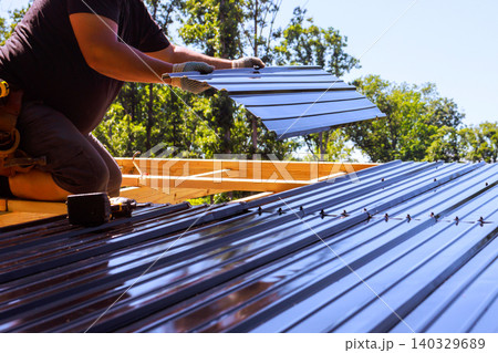 Contractor roofer installs metal roofing panels on wooden beams frame at construction site surrounded by trees. 140329689