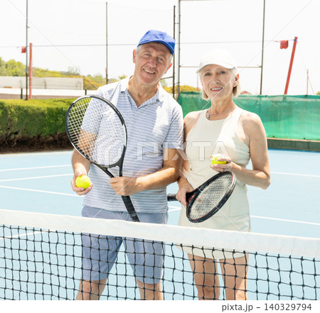 Positive old man and woman in sports clothes posing with racket in hand next to net in open air tennis court 140329794