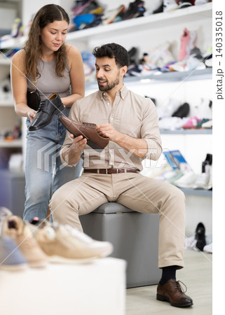 Man tries on classic shoes while sitting in a store, next to him stands a woman who helps Man tries on classic shoes while sitting in a store, next to him stands a woman who helps 140335018