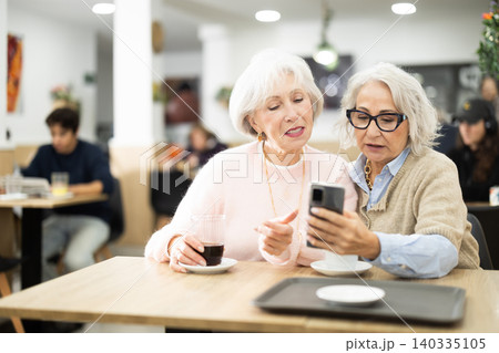 Two senior women colleagues meeting and drinking coffee at table in cafeteria, looking at photos on mobile phone Two senior women colleagues meeting and drinking coffee at table in cafeteria, looking at photos on mobile phone 140335105