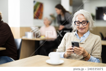 European elderly woman drinking fresh coffee at table in cafe and looking at mobile phone 140335871